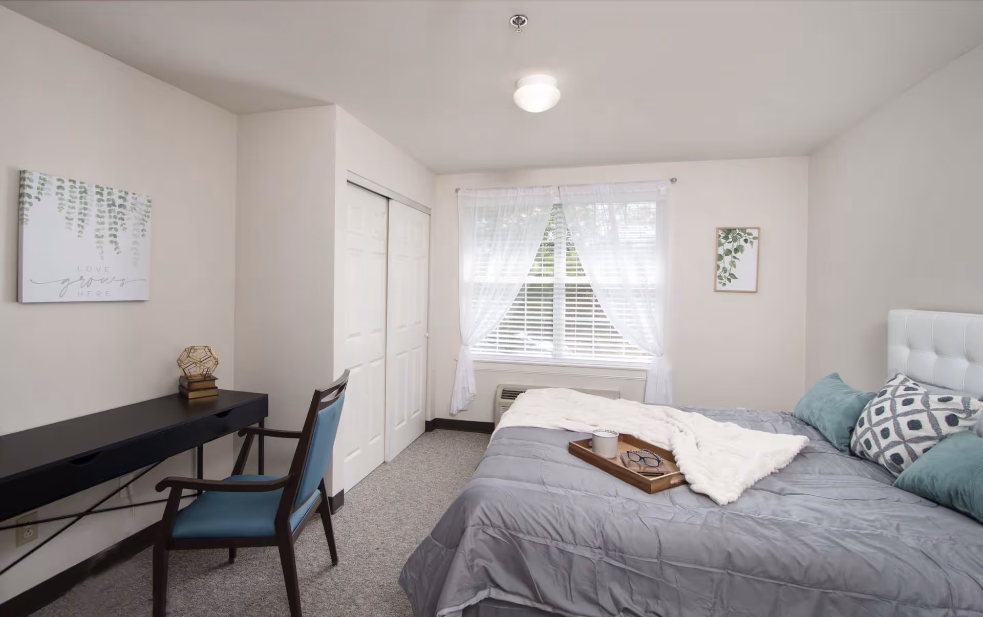 A bright bedroom with a large bed covered in a gray comforter and decorative pillows. A wooden tray with a cup and glasses rests on the bed. There is a window with sheer white curtains letting in natural light. A black desk with a blue chair is positioned against one wall, with a decorative item and books on the desk. Two framed botanical prints hang on the walls. A closet with white sliding doors is visible next to the window.