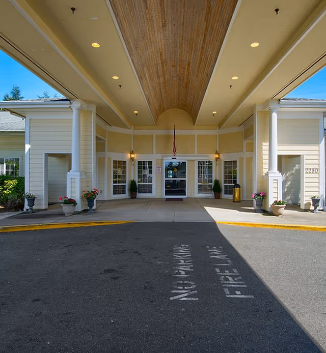 Entrance area of Inland Point Retirement Community featuring a covered drop-off zone with white columns, potted plants, and a glass door entrance. The ceiling has recessed lighting and a wooden panel in the center. The pavement has markings indicating 'NO PARKING FIRE LANE'.
