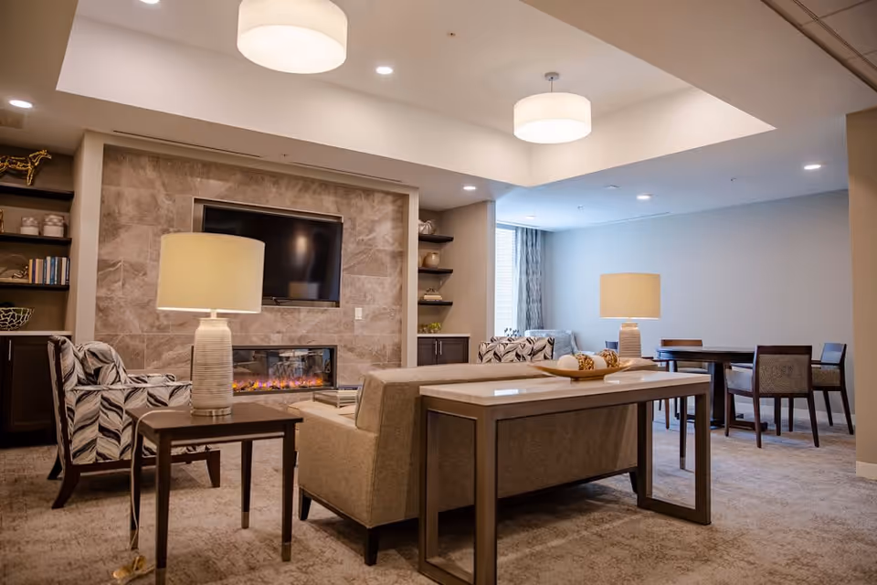 A cozy living room area with a beige sofa, patterned armchairs, two table lamps, a wall-mounted flat-screen TV above an electric fireplace, and a dining table with chairs in the background. The room features soft lighting and neutral tones.