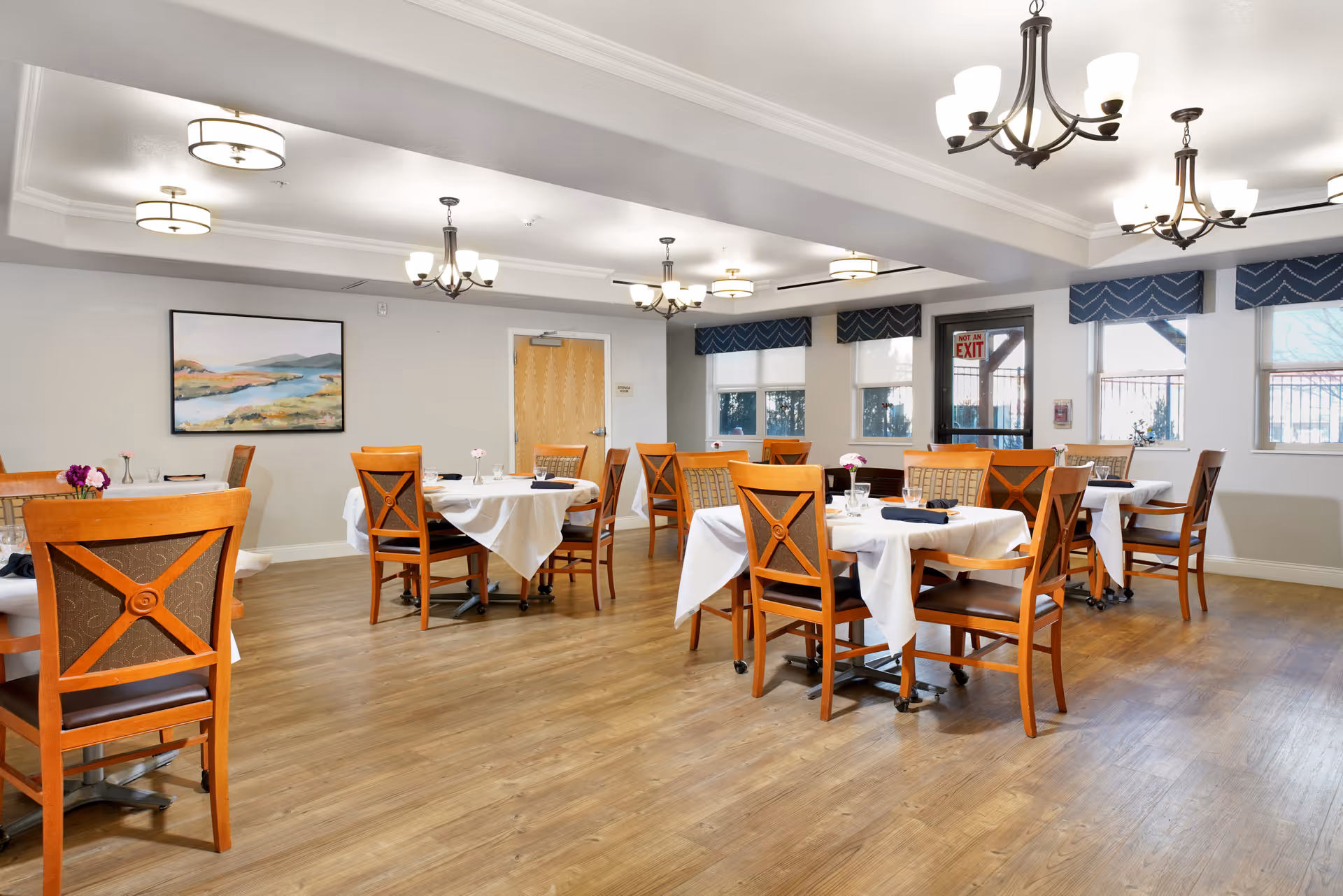 A bright dining room with wooden tables and chairs set with white tablecloths and overhead chandeliers.