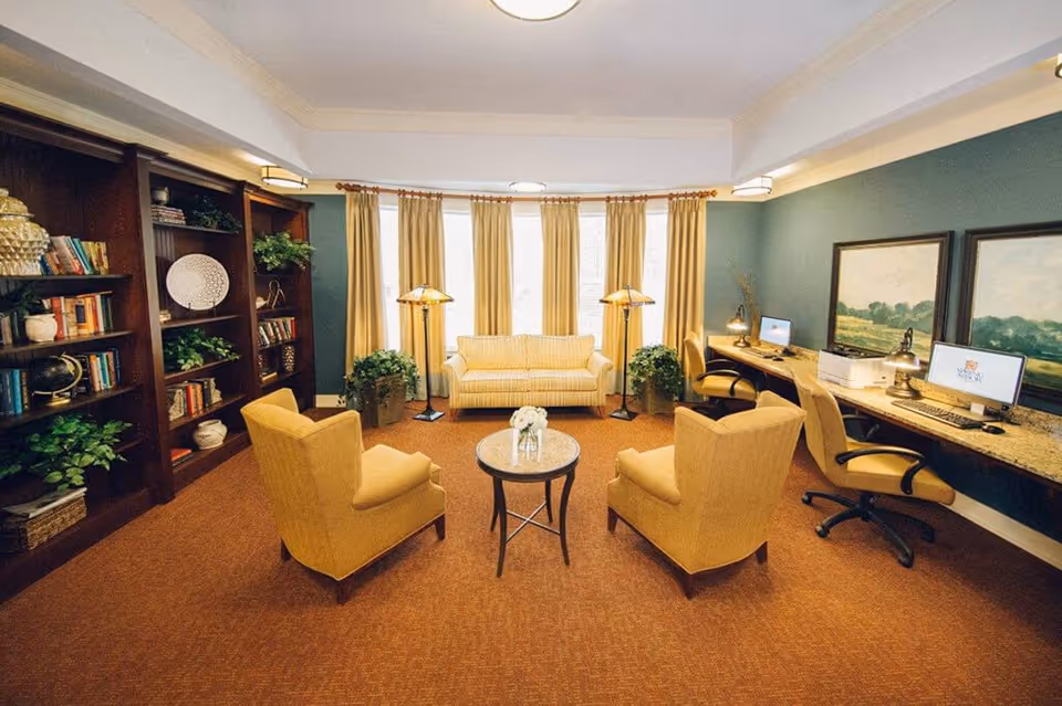 A cozy living room area with a beige sofa, two matching armchairs, a small round table with a flower vase, and two floor lamps near large windows with beige curtains. To the left, there is a wooden bookshelf filled with books and decorative items. On the right side, there is a long desk with three office chairs, two computer monitors, a printer, and two framed landscape paintings on the wall.