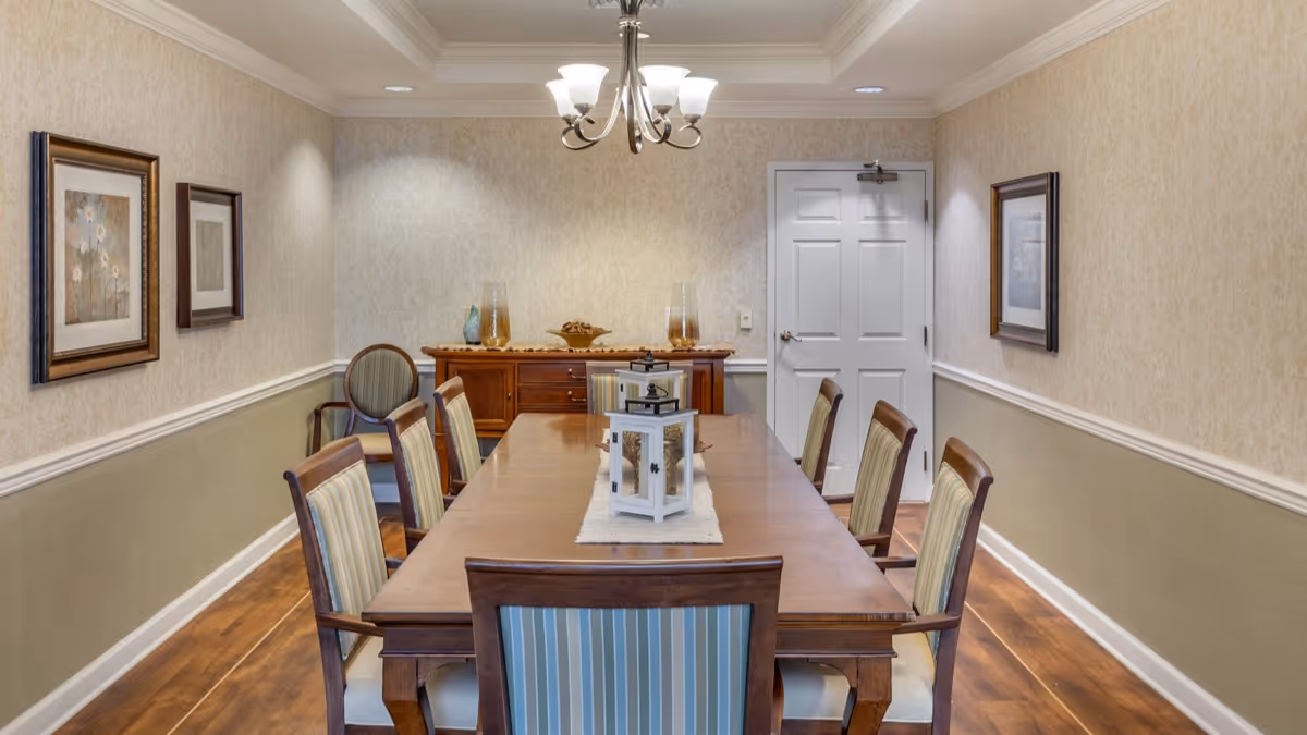 A formal dining room with a long wooden table surrounded by eight striped upholstered chairs. The walls are decorated with framed artwork and have a two-tone paint design with a chair rail. A sideboard with decorative items is against the far wall beneath a ceiling light fixture.