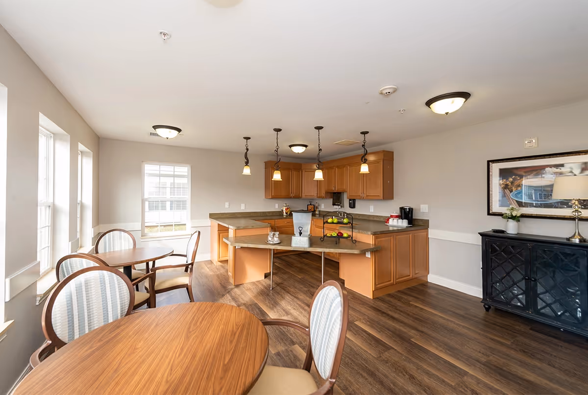 A bright and clean dining area with round wooden tables and cushioned chairs near large windows. In the background, there is a kitchen area with wooden cabinets, a countertop with a water dispenser, fruit stand, and coffee maker. The room has wood flooring, ceiling lights, and a framed picture on the wall above a dark cabinet with a lamp and small plant.