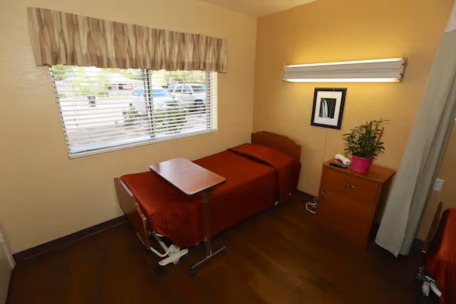 A small room with a single bed covered in a red blanket, a wooden bedside table with a potted plant and a telephone, a wall-mounted light fixture above a framed picture, and a window with blinds and a beige valance. The floor is wooden and there is a movable overbed table positioned over the bed.