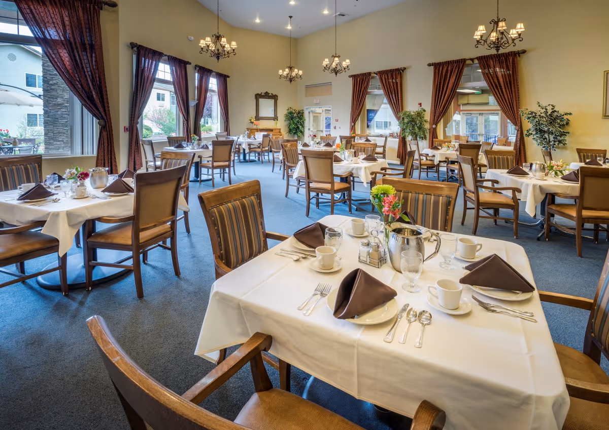 A spacious dining room with multiple tables covered in white tablecloths, each set with brown napkins, cups, glasses, silverware, and small flower arrangements. The room has large windows with brown curtains, blue carpet, and chandeliers hanging from the ceiling. There are wooden chairs with striped cushions around the tables.