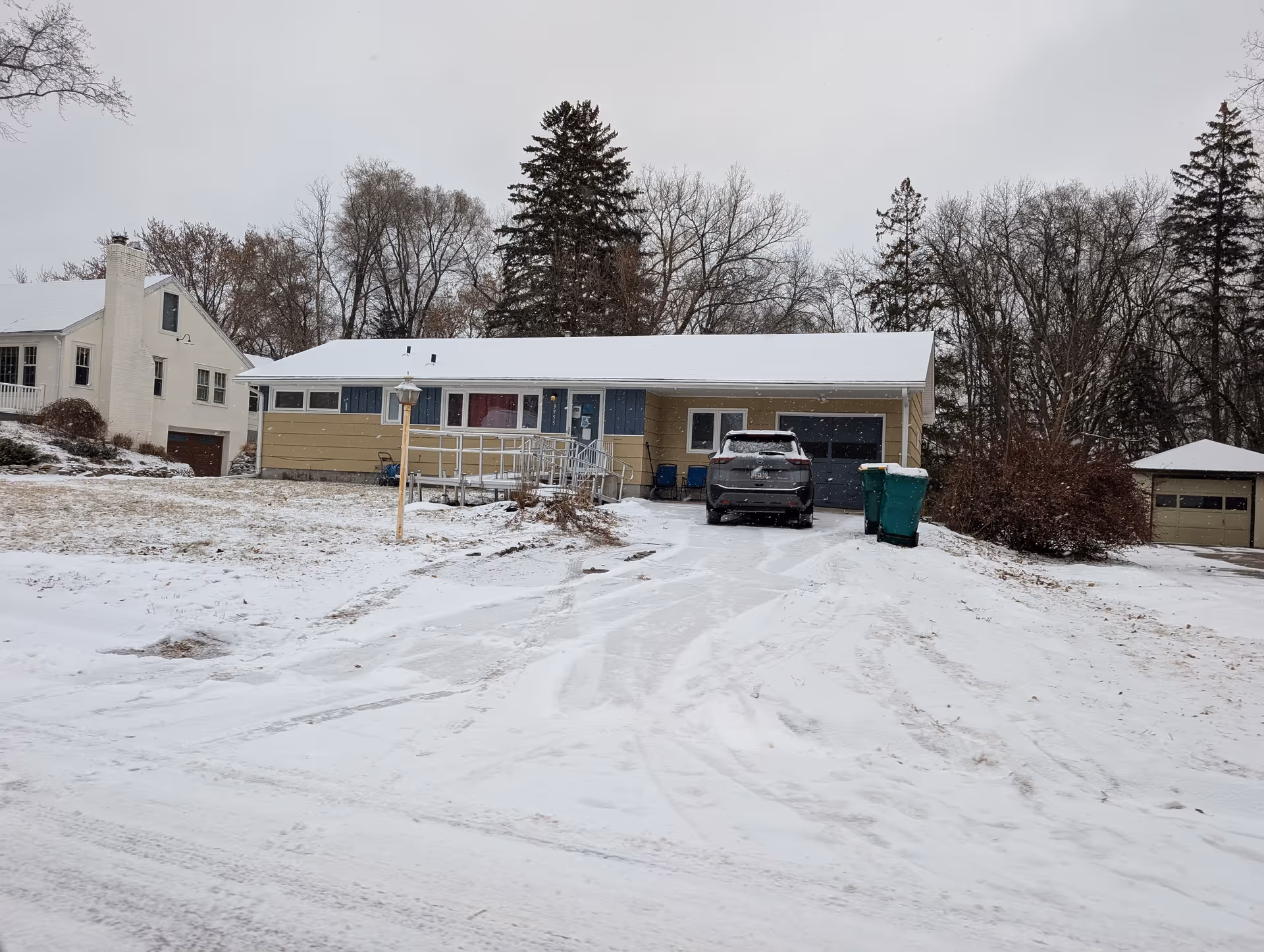 Single-story ranch house with a car in the snowy driveway, a front wheelchair ramp, and trees in the background.