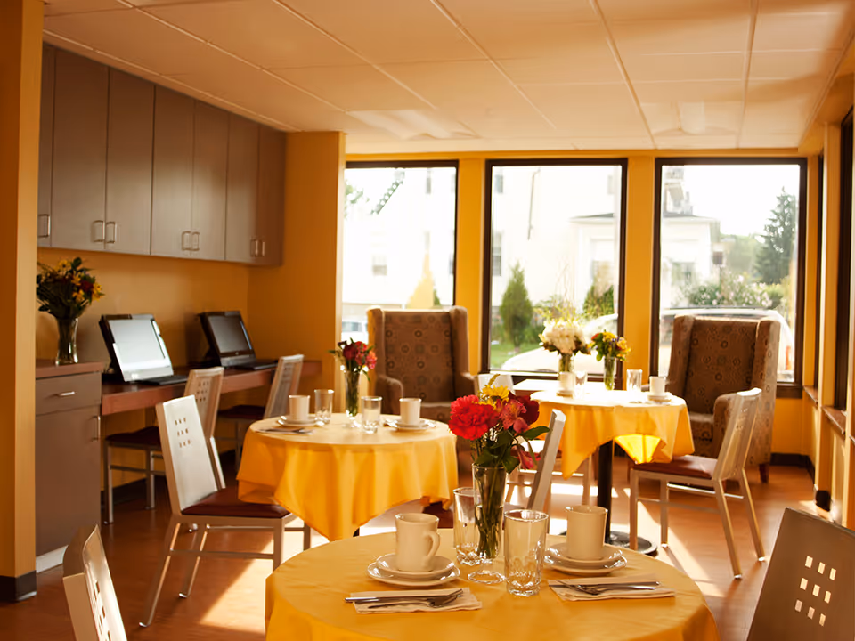 A bright dining room with round tables covered in yellow tablecloths, each set with white cups, saucers, glasses, and silverware. Vases with colorful flowers are placed on the tables. There are cushioned armchairs and regular chairs around the tables. Large windows let in natural light, and a counter with computers and chairs is visible along one wall.