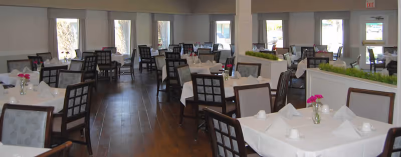 A spacious dining room with multiple tables covered in white tablecloths, each set with napkins, cups, and small vases containing pink flowers. The room has wooden flooring and large windows allowing natural light to brighten the space.
