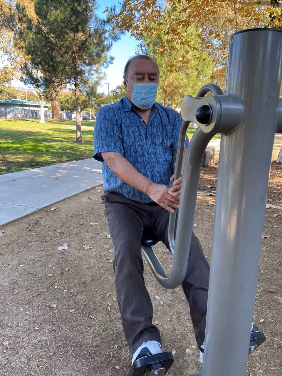 An elderly man wearing a blue patterned shirt, dark pants, white sneakers, and a face mask is using outdoor exercise equipment in a park with trees and a paved walkway nearby.