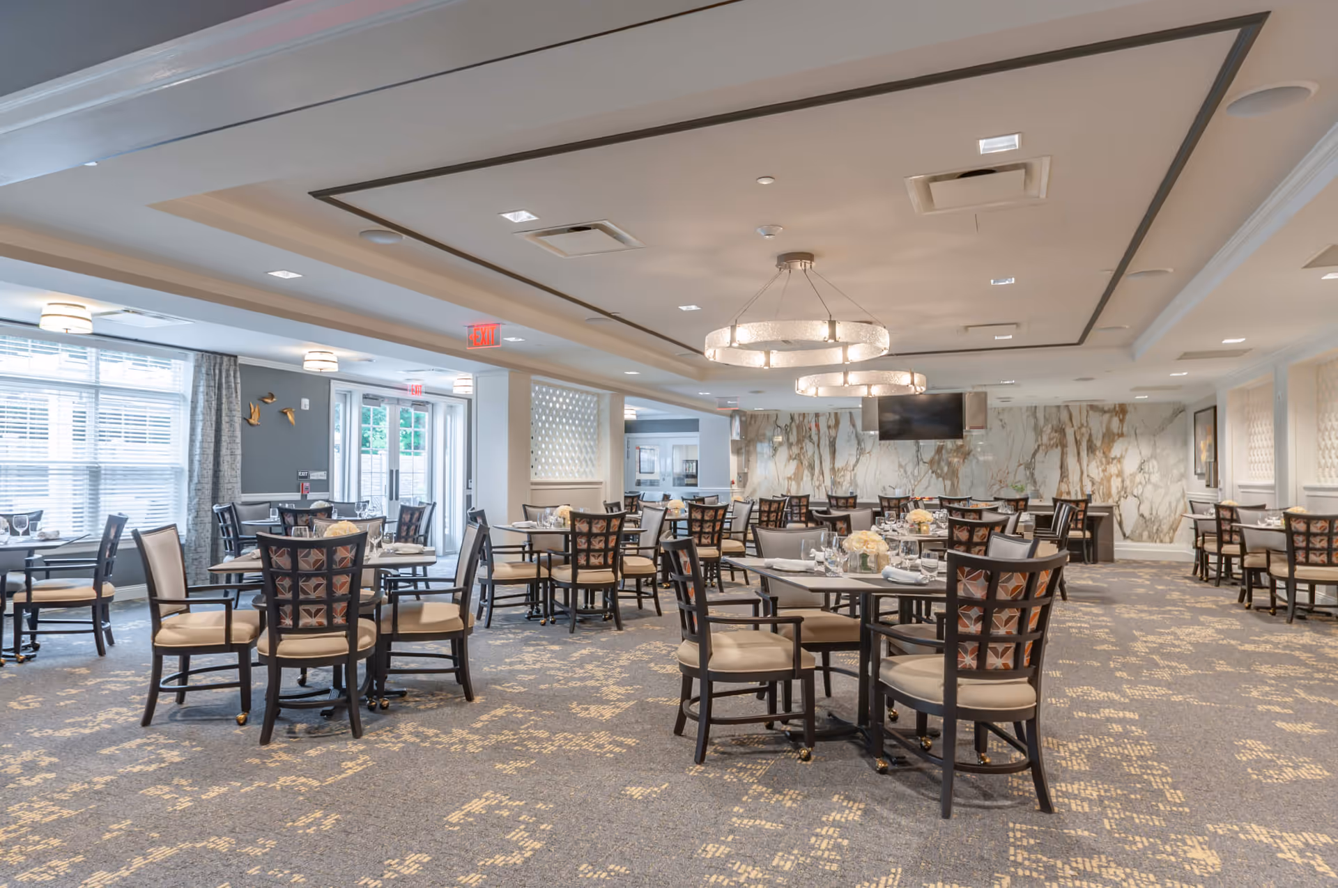 A spacious dining room with multiple round tables and cushioned chairs arranged neatly. The room features large windows with curtains, modern circular chandeliers, a patterned carpet, and a marble accent wall with a mounted television.