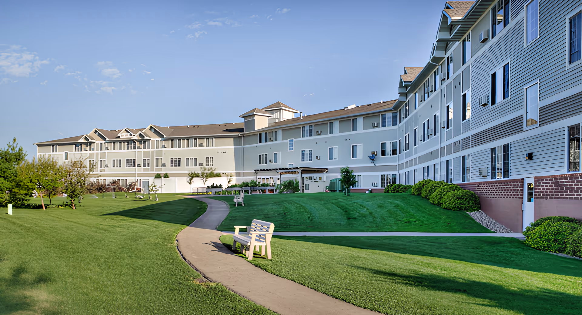 Multi-story senior living building overlooking a curved pathway, manicured lawn, and benches under a blue sky.