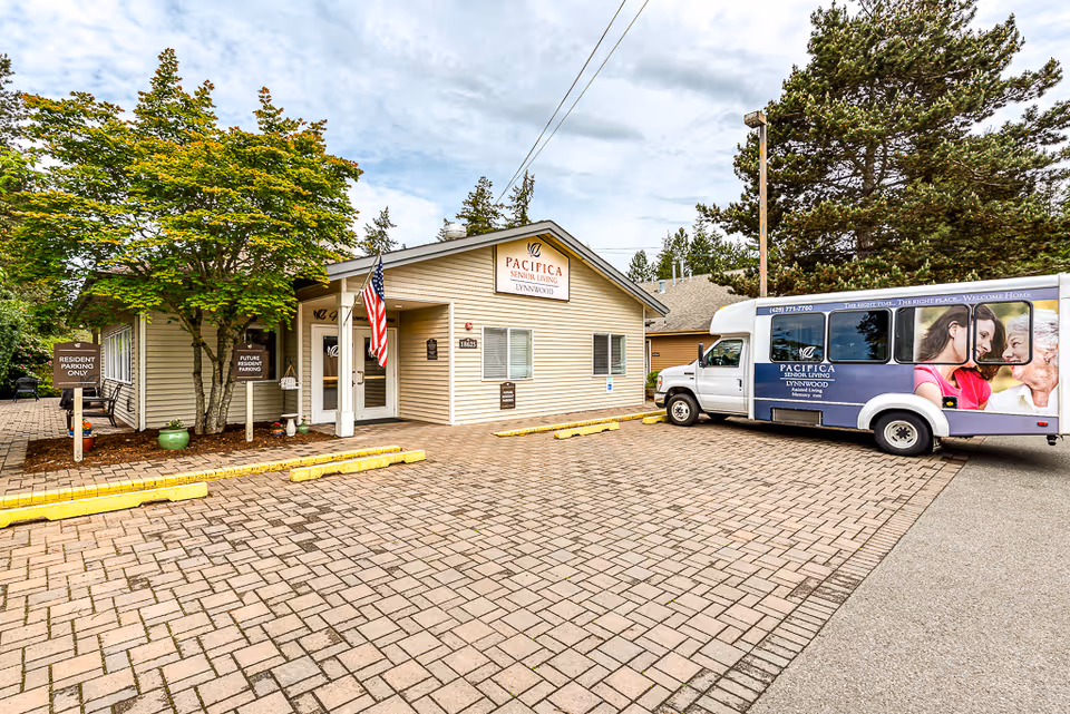Exterior view of a senior living facility building with a beige exterior, an American flag near the entrance, and a Pacifica Senior Living Lynnwood shuttle bus parked on the right. There are trees and signs indicating resident and future resident parking.