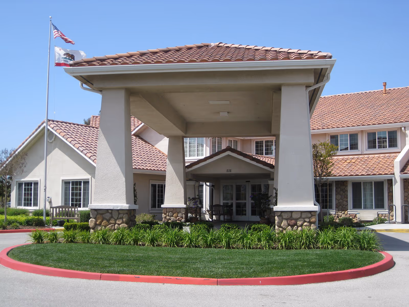 Front entrance of a senior living facility with a covered driveway supported by large pillars with stone bases. The building has a tiled roof and multiple windows. Two flags, one American and one California state flag, are flying on a flagpole to the left. There is a circular landscaped area with green grass and plants in front of the entrance.