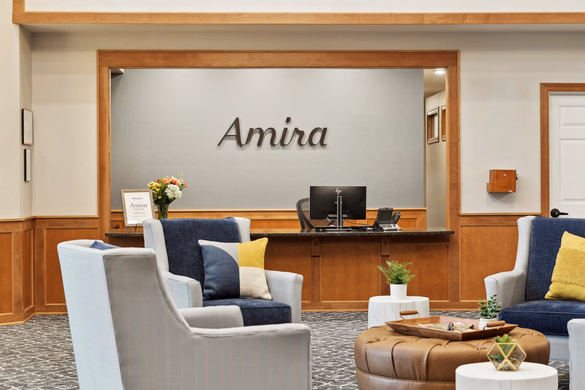 Reception area of Amira Choice Forest Lake featuring a wooden desk with a computer monitor, a vase of flowers, and a welcome sign. In front of the desk are comfortable armchairs with blue and yellow cushions, a round brown ottoman with a tray, and small white tables with plants. The wall behind the desk has the word 'Amira' mounted on it.