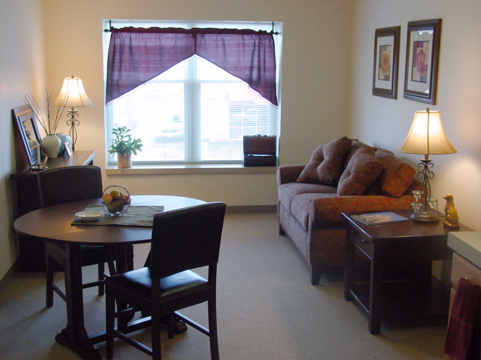 A cozy living area with a small round dining table and two chairs on the left, a window with purple curtains in the center, and a patterned sofa with multiple cushions on the right. There are two table lamps on either side of the room, a side table with a magazine, and framed floral artwork on the wall above the sofa.