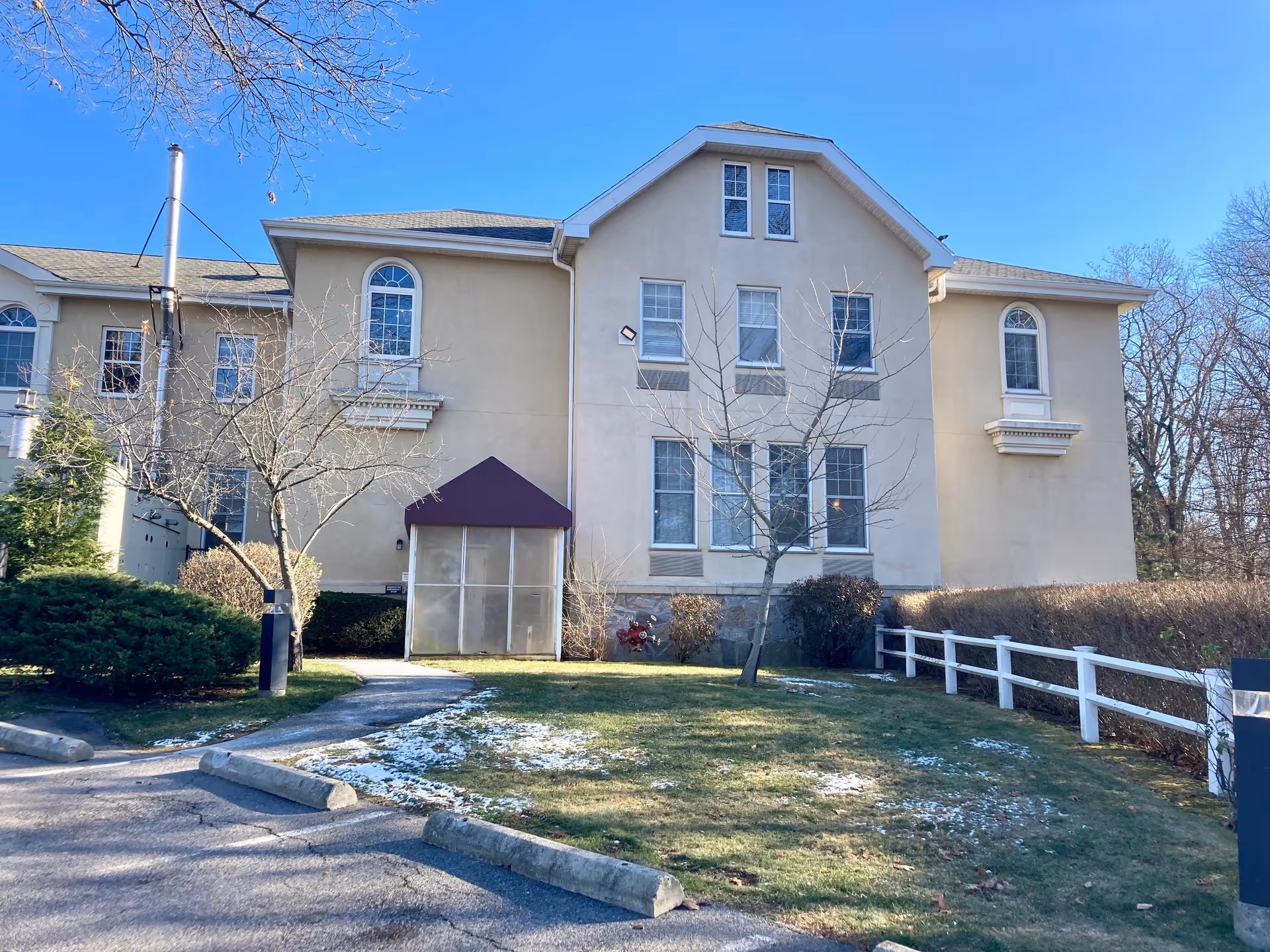 Exterior view of a beige two-story building with multiple windows, a small entrance with a maroon awning, leafless trees, and a grassy area with patches of snow in front. The sky is clear and blue.