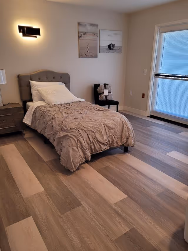 A neatly made single bed with a tufted headboard in a room with wood-patterned flooring. There is a nightstand with a lamp to the left of the bed, two framed pictures on the wall above a black chair with folded blankets, and a large window with blinds on the right side.