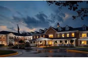 Exterior view of The Gardens at Fair Oaks facility at dusk, showing a large two-story building with multiple windows lit from inside, a covered entrance with columns, and a circular driveway with landscaping and trees under a cloudy sky.