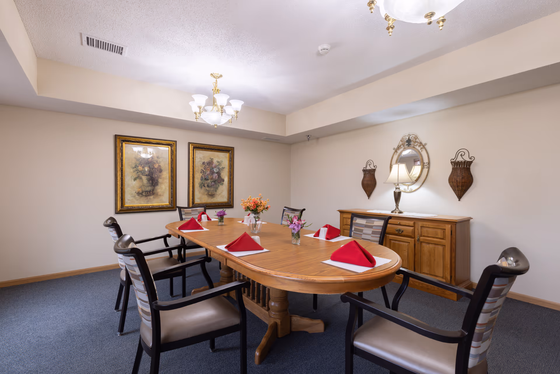 A dining room with a wooden oval table set with red folded napkins on white placemats, surrounded by six chairs with cushioned seats and backs. The room has beige walls, two framed floral paintings, a wooden sideboard with a lamp and a round mirror above it, and two decorative wall hangings. A chandelier with multiple lights hangs from the ceiling.