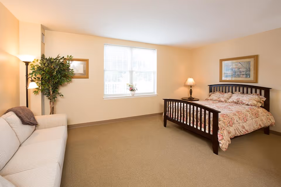A bright bedroom with beige walls and carpet, featuring a dark wooden bed with floral bedding, a matching nightstand with a lamp, a large window with white blinds, a beige striped sofa with a throw blanket, a tall floor lamp, a potted plant, and framed artwork on the walls.