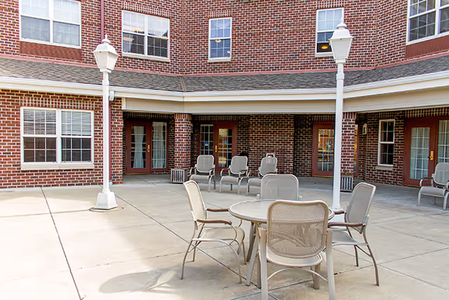 Outdoor patio area at a senior living facility with a round table and several chairs arranged around it. The patio is surrounded by a brick building with multiple windows and doors. Two white lamp posts are visible on the patio.