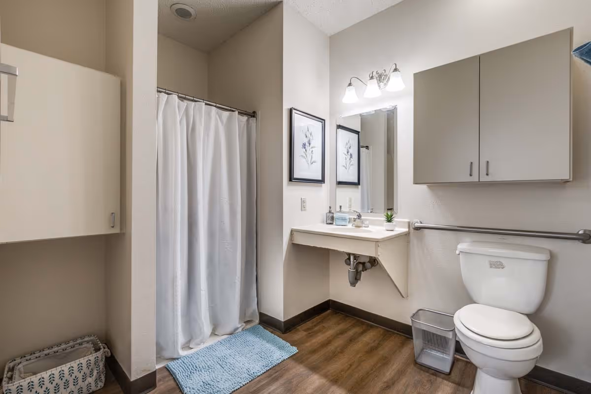 A clean and simple bathroom with a white shower curtain, a blue bath mat, a wall-mounted sink with a mirror and three light fixtures above it, a toilet with a grab bar next to it, a small trash bin, and wall cabinets for storage. The floor has a wood-like finish.