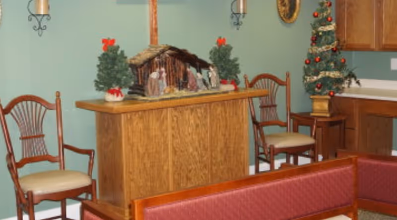 Small interior common room with wooden chairs and benches, a nativity scene and small decorated Christmas trees on a wooden counter against a green wall.