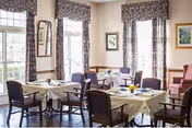A bright dining room with round tables, chairs, and floral curtains along windows.