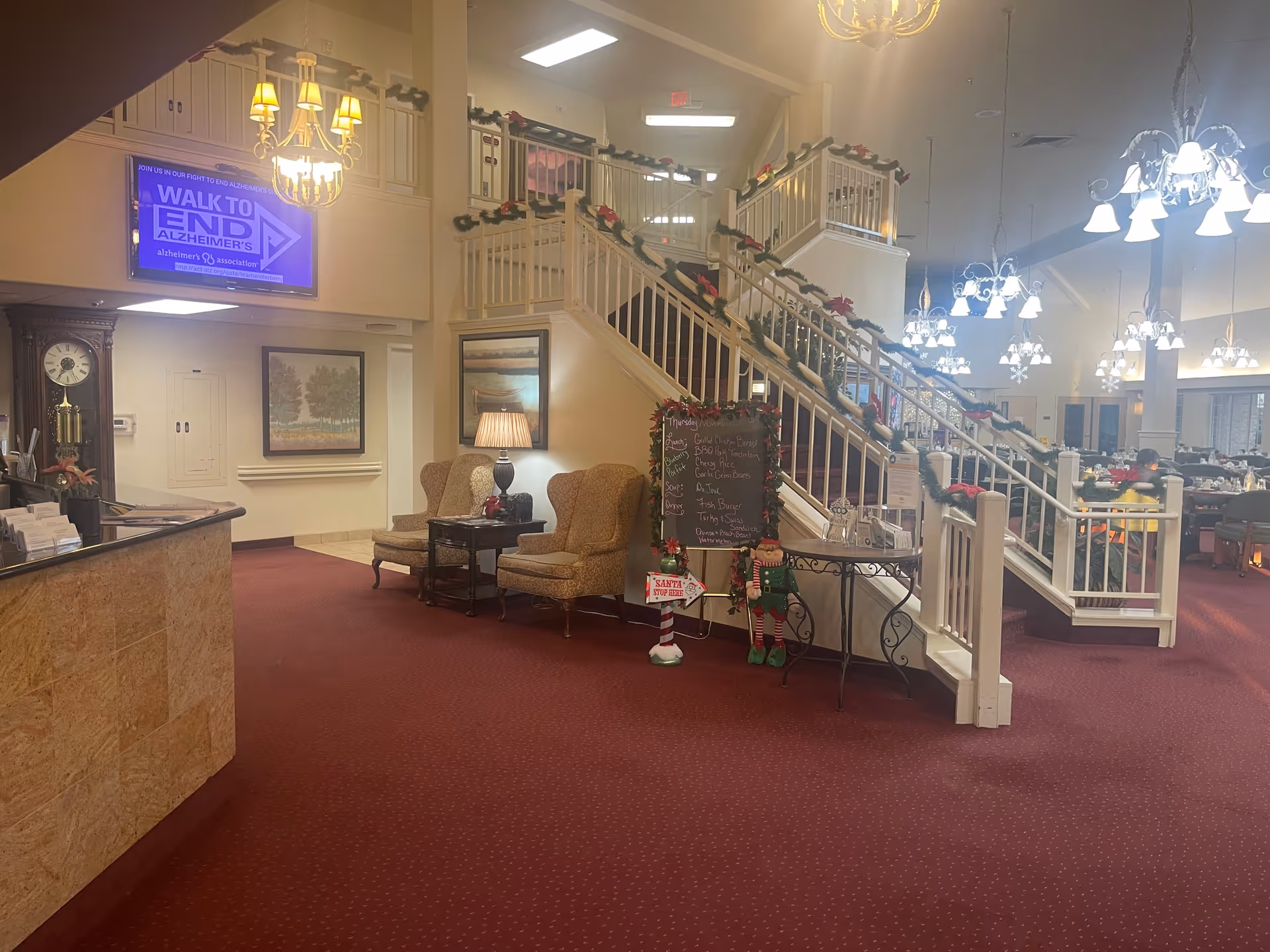 Interior view of a senior living facility lobby area with a red carpet, two upholstered chairs with a small table and lamp between them, a staircase decorated with garlands and poinsettias, a chalkboard menu, and a dining area with tables and chairs visible in the background. A large clock and framed paintings are on the walls, and multiple chandeliers provide lighting.