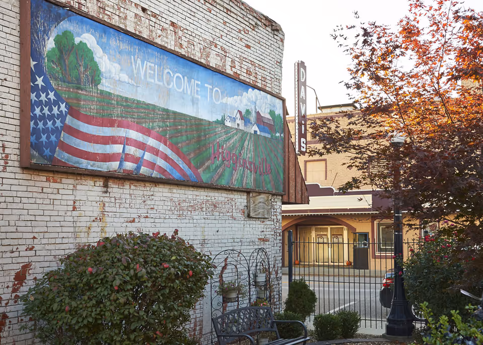 Brick building exterior with a large painted 'Welcome to' mural, a bench, shrubs, a street-facing gate and neighboring storefronts.