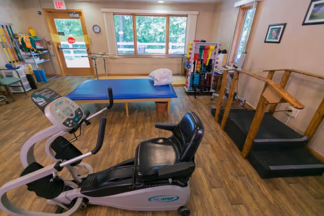 A rehabilitation room with exercise equipment including a NuStep recumbent cross trainer, a blue therapy table with a pillow, parallel bars for walking exercises, and various physical therapy tools organized on racks. The room has wooden flooring, large windows letting in natural light, and an exit door with a stop sign.