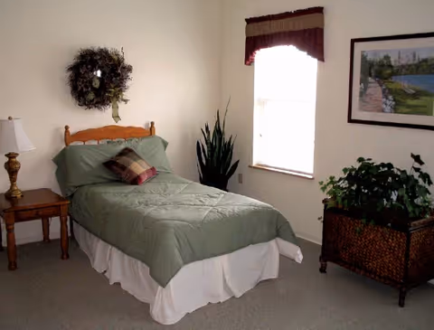 A simple bedroom with a single bed covered in a green comforter and a plaid pillow. There is a wooden nightstand with a lamp on the left side of the bed, a decorative wreath hanging on the wall above the headboard, a tall green plant near the window, and a framed picture on the wall to the right. A woven basket with a large green plant is placed under the picture.