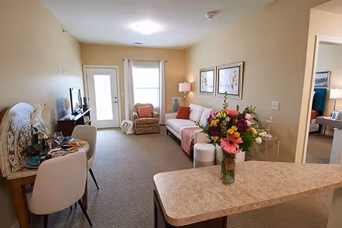 A cozy living room area in a senior living facility with a beige sofa, a floral armchair, a small dining table with two chairs, a TV on a stand, and a countertop with a vase of colorful flowers. The room has beige walls, carpeted floor, framed artwork on the wall, and a door with a window letting in natural light.