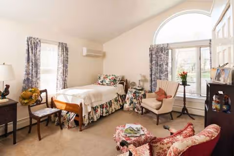 Bright senior living bedroom with floral bedspread, arched window, and seating area.