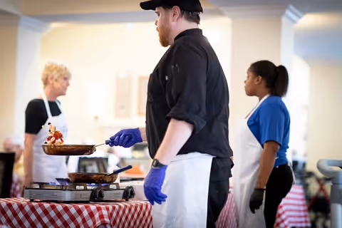 Three staff in aprons and gloves cook and serve food on portable burners atop a red-checkered tablecloth in a dining area.