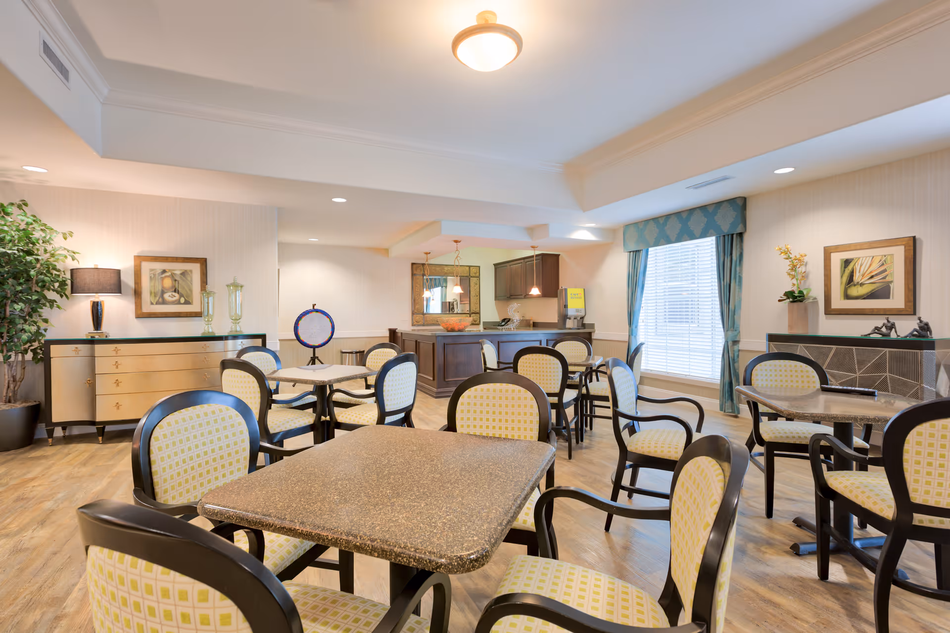 A bright and spacious dining room in a senior living facility with multiple tables and chairs arranged neatly. The chairs have a light yellow patterned upholstery with dark wooden frames. The room features wood flooring, a large window with blue patterned curtains, decorative plants, framed artwork on the walls, and a buffet or serving area in the background with a water dispenser and pendant lights overhead.
