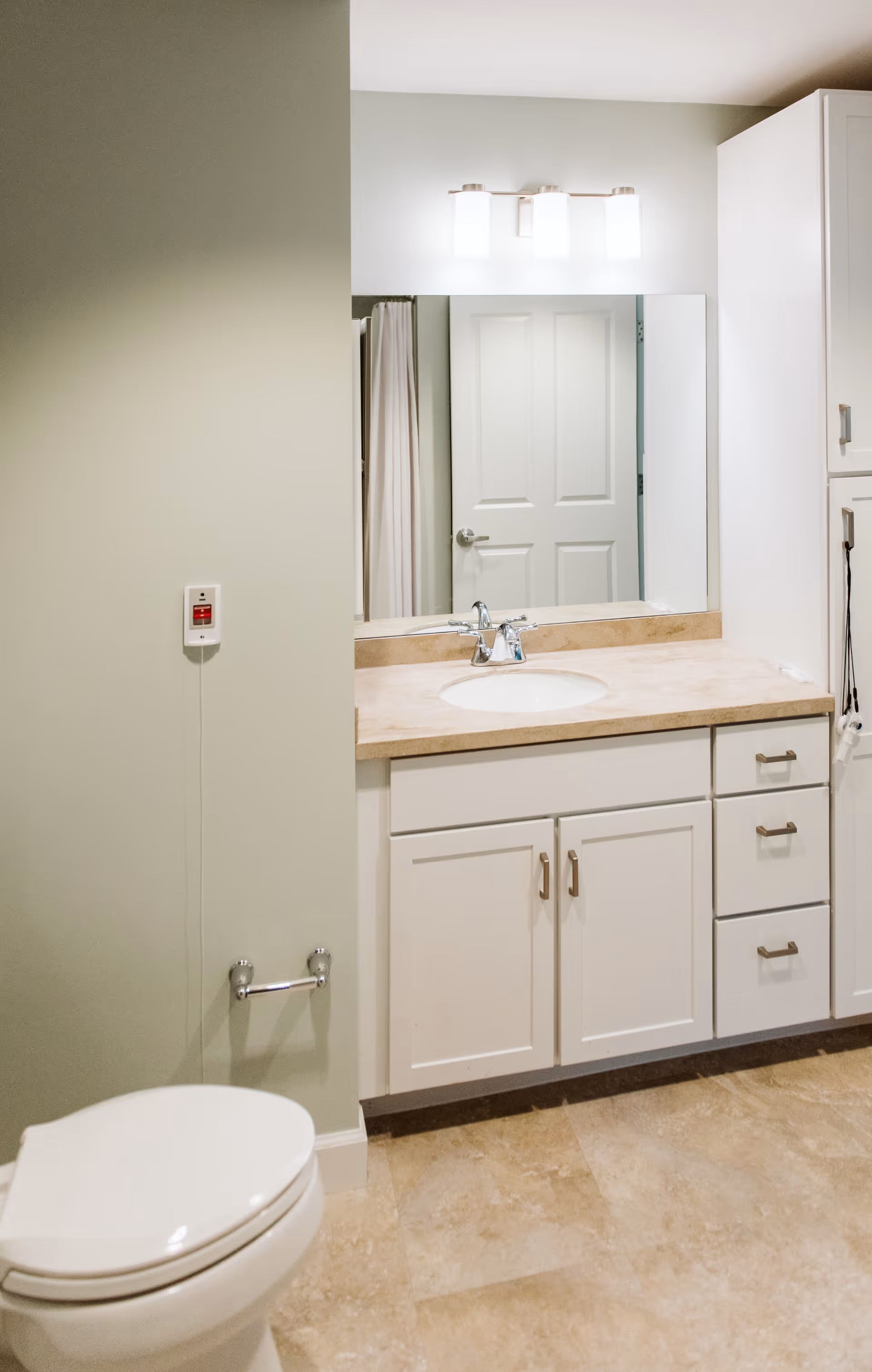 A clean bathroom featuring a white toilet, a beige countertop with an under-mount sink, white cabinetry with drawers and doors, a large mirror above the sink, a three-light fixture above the mirror, and a closed white door reflected in the mirror. The floor is tiled in a beige color.
