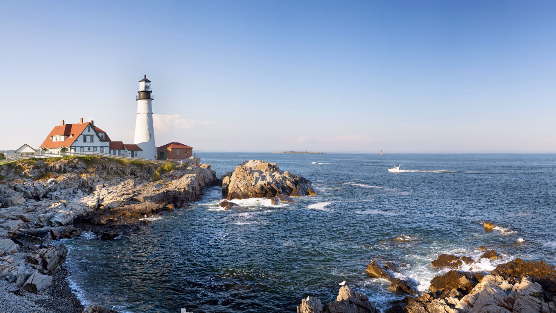 A lighthouse and adjacent buildings on a rocky coastline overlooking the ocean with a small boat in the distance.