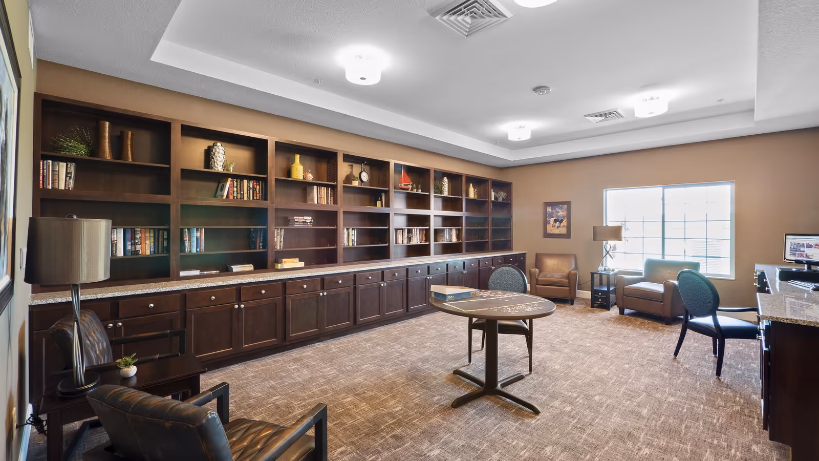 A spacious room with a long wooden bookshelf filled with books and decorative items along one wall. The room has beige walls and carpeted floor. There are several chairs including leather armchairs and cushioned chairs around a small round table with a puzzle on it. A large window lets in natural light, and there are ceiling lights and a floor lamp providing additional lighting. A computer workstation is visible on the right side of the room.