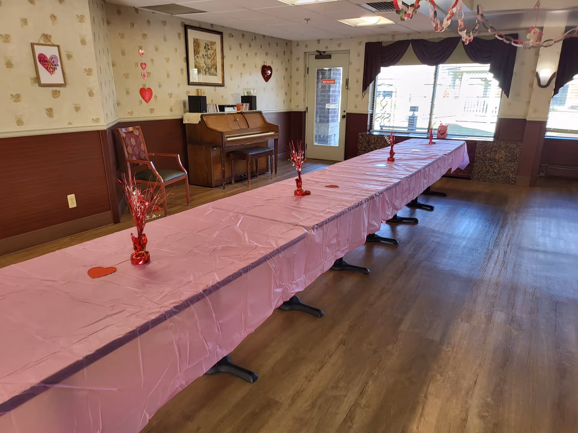 A long table covered with a pink tablecloth and heart-themed centerpieces set up in a senior living dining/activity room with a piano and large windows.