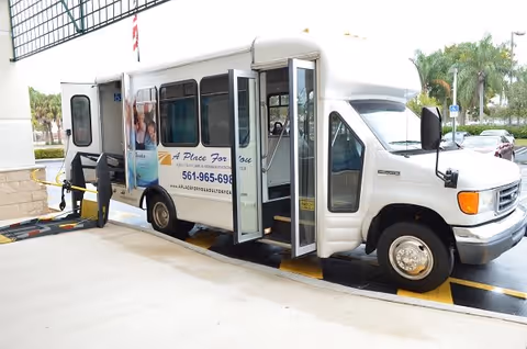 A white shuttle bus with the name 'A Place For You Adult Daycare and Rehab Center' parked outside a building. The bus door is open, and there is a wheelchair lift extended on the side. Palm trees and parked cars are visible in the background.