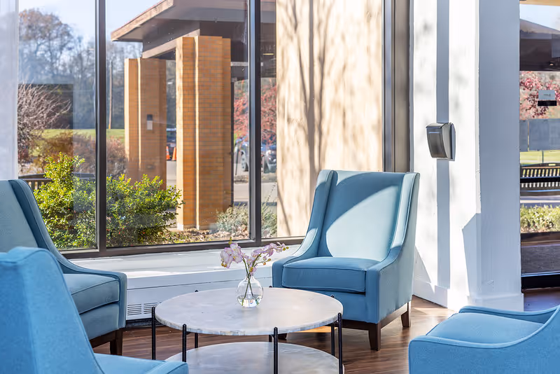 A bright seating area with three blue upholstered armchairs arranged around a round marble coffee table with a small vase of flowers. Large windows behind the chairs show an outdoor view with brick pillars, greenery, and trees.