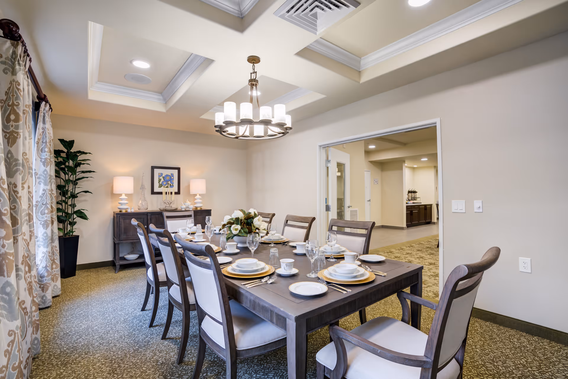 Dining room in a senior living facility with a long table set for a meal, chandelier overhead, a sideboard with lamps, and patterned curtains.