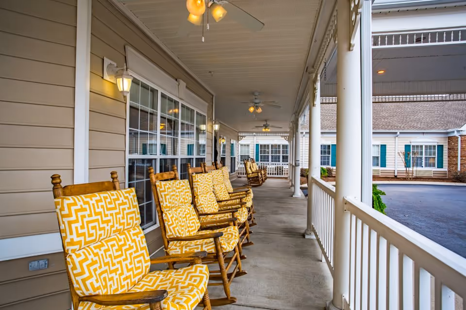 Covered front porch at a senior living facility with a row of yellow-patterned cushioned rocking chairs along a white railing.