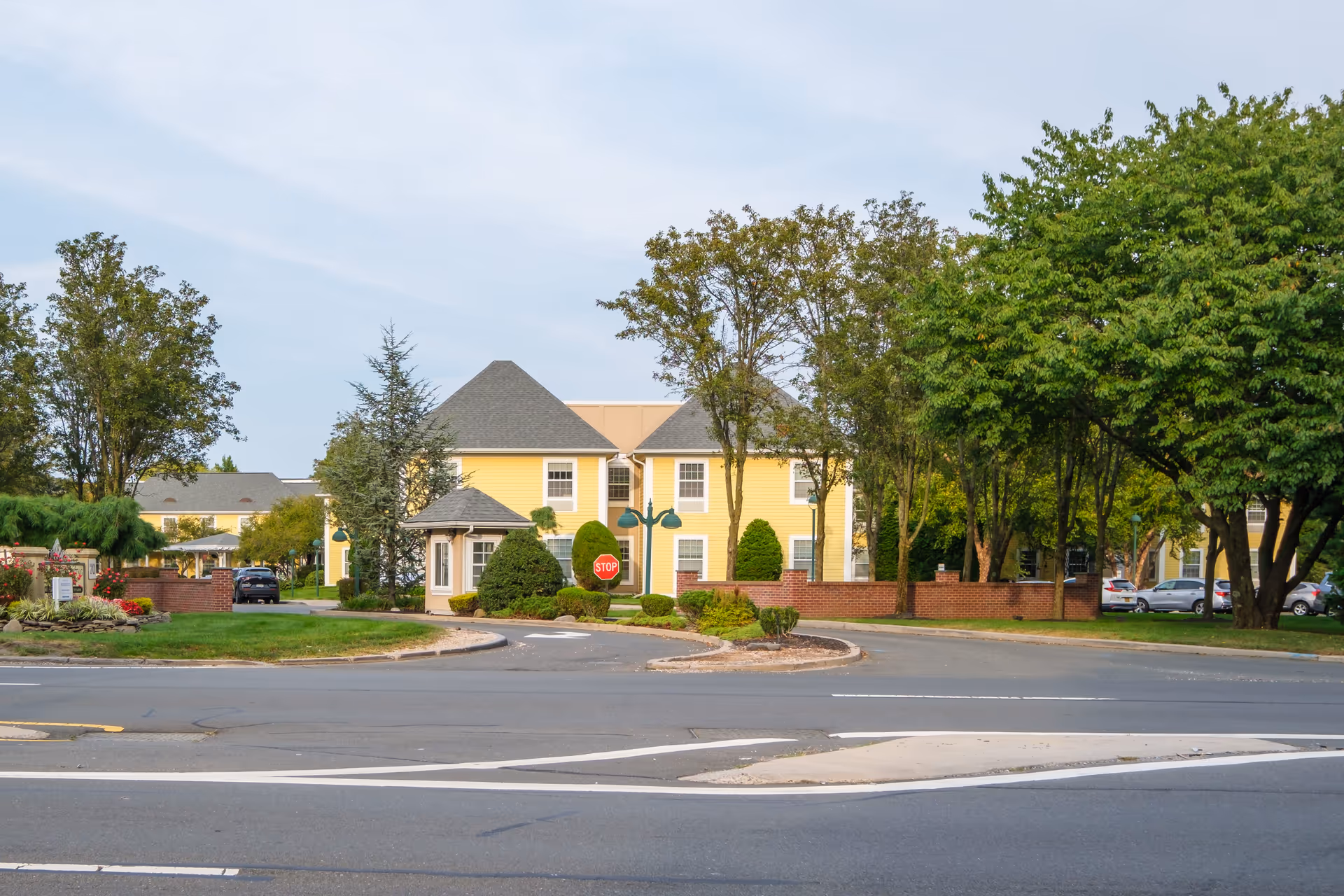View of the exterior of a senior living facility with yellow buildings, a small guardhouse, trees, and a stop sign near the entrance. Several cars are parked in the background.
