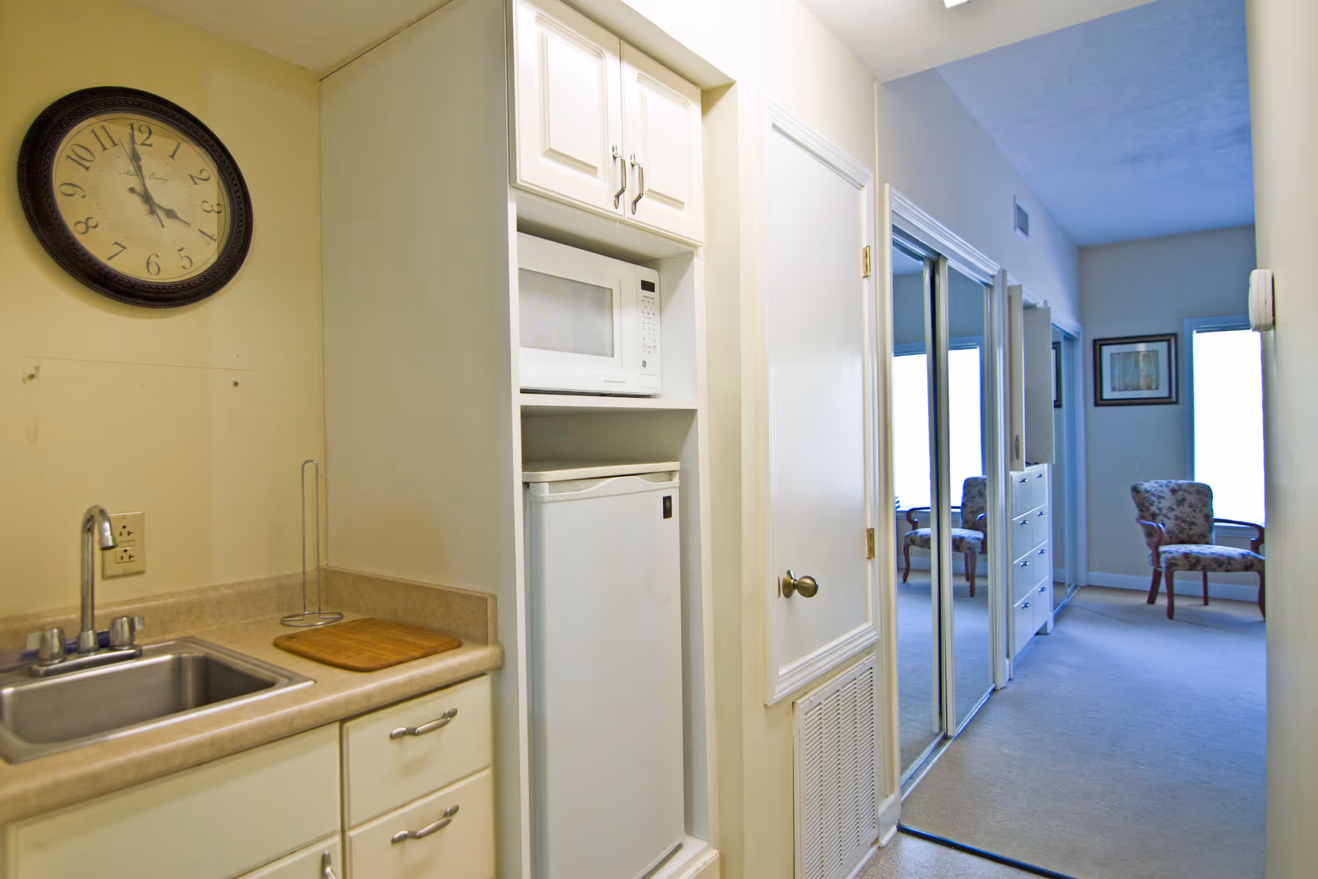 Interior view of a small kitchenette area with a sink, countertop, microwave, and mini refrigerator. A wall clock is mounted above the sink. The kitchenette opens into a hallway with mirrored closet doors and a floral upholstered chair near a window at the end.
