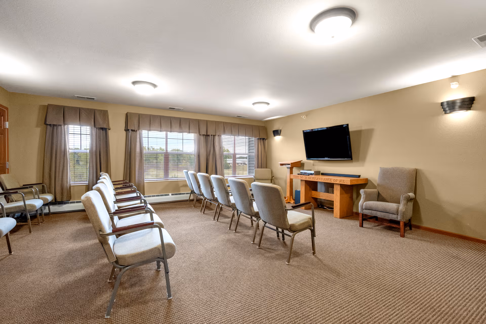 A meeting or activity room in a senior living facility with two rows of chairs facing a wall-mounted flat screen TV. The room has beige walls, carpeted floor, large windows with curtains, and a wooden podium and table beneath the TV.