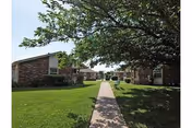 Concrete walkway through green lawns and shade trees between single-story brick apartment buildings.
