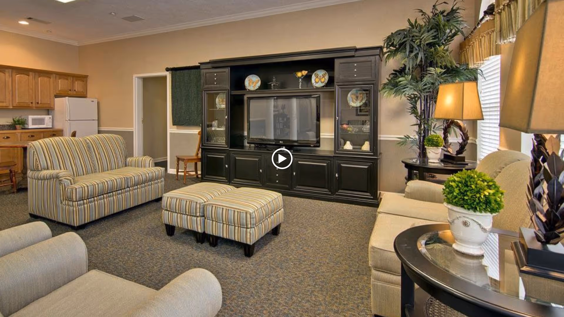 Communal living room with striped sofas and ottoman facing a large black entertainment center, with side tables, lamps, and plants.