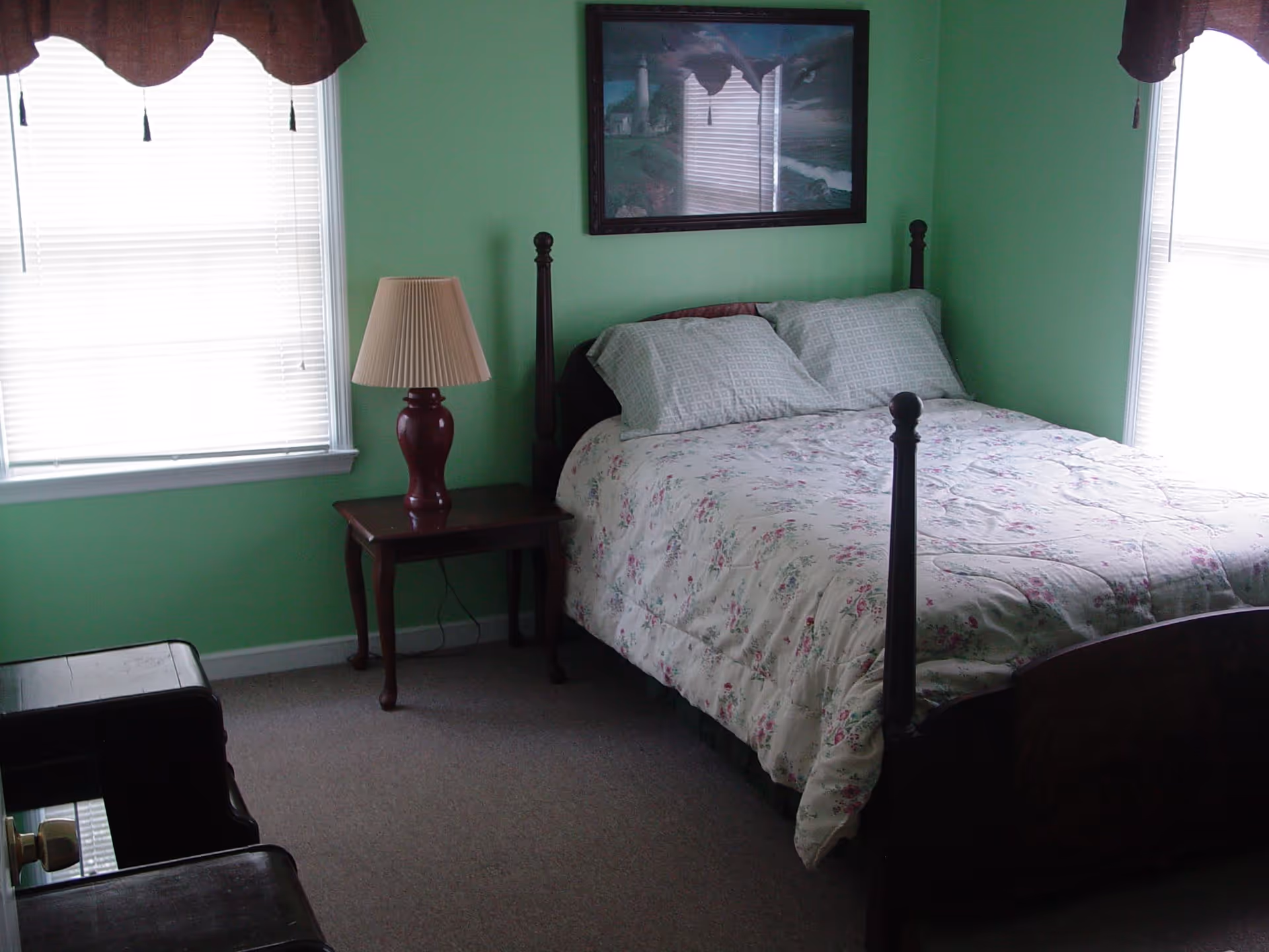 A bedroom with light green walls, a bed with floral bedding and two pillows, a wooden nightstand with a red lamp, two windows with white blinds and brown valances, and a framed picture hanging above the bed.